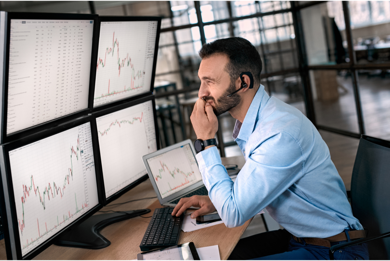 Man in a blue shirt working at a desk with multiple computer monitors displaying financial charts.